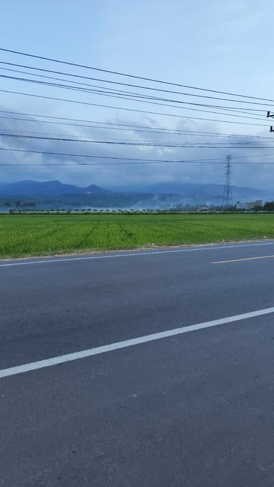 The image depicts a landscape in Bateiiliek, Indonesia. In the foreground, there is an asphalt road with a white lane marking. Adjacent to the road is a vibrant green rice field. In the background, there are mountains partially obscured by clouds. Overhead, power lines span the sky. An electrical transmission tower is visible on the right side of the background. The overall scene suggests a rural setting with agricultural land near infrastructure.