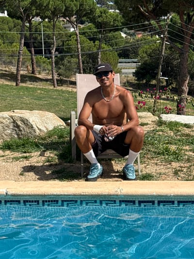 A man is sitting on a lounge chair by a swimming pool. He is shirtless, wearing black shorts, white socks, and blue sneakers. He has a black baseball cap and sunglasses on and is wearing a silver chain. The man is holding a clear cup. The lounge chair is beige and is on a paved area next to the pool. Behind the man is a grassy area with trees and rocks. The swimming pool has blue water with white lines on the bottom and a tiled edge. The scene appears to be a backyard or resort in Sant Vicenç de Montalt, Spain.