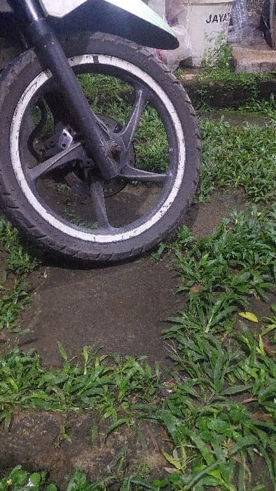 The image shows the front tire of a motorcycle parked on a dirt path. The tire has a white rim and is partially obscured by green foliage. The motorcycle is parked near a concrete wall with the word "JAYAN" written on it. The scene is located in Kecamatan Babakan Madang, Indonesia, at the coordinates -6.579 latitude and 106.886 longitude. There are no people or other objects visible in the image.