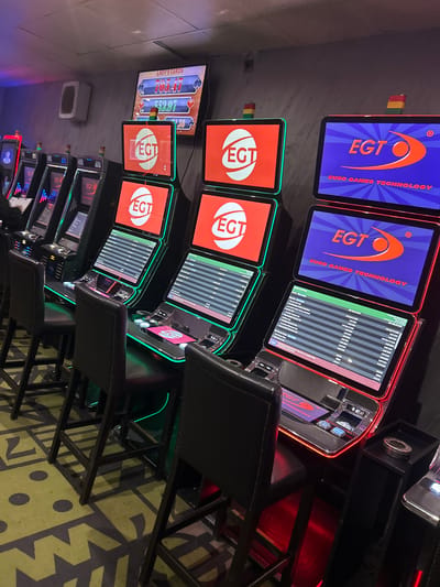 The image shows a row of slot machines inside what appears to be a casino. The machines are from "Euro Games Technology" (EGT). Some screens display the EGT logo, others show the game "Lady's Cards" with various amounts displayed, and some screens display lists of payout information in EUR. The chairs in front of the machines are black leather with metal frames. The lighting is dim, creating a typical casino atmosphere. The floor covering shows an abstract pattern in shades of green and black. The setting implies an indoor gambling establishment, with no direct visual cues linking it to Krivodol, Bulgaria, or any other specific location.