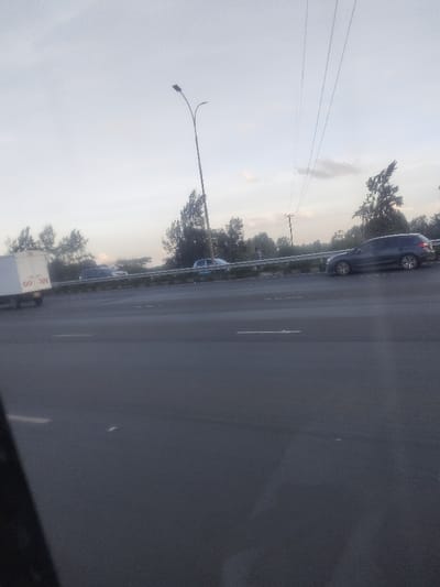 The image depicts an outdoor scene on a multi-lane highway in Theta ward, Kenya, under a pale, overcast sky. The perspective is from within a vehicle, with a blurry dark object partially obscuring the lower left foreground.

The foreground consists of dark asphalt pavement with indistinct white lane markings. A metal guardrail separates the immediate lanes from an opposing carriageway. Behind this guardrail, a dense strip of green and brown foliage, composed of bushes and trees, runs horizontally across the midground.

Vehicles are present on the opposite side of the road. From left to right, a white box truck with visible red/orange chevron markings on its rear is partially obscured. Further right, a light-colored sedan or small SUV is discernible. To the far right, a dark-colored SUV is positioned near the roadside, appearing stationary or moving slowly.

In the background, a tall, brown street light pole with two light fixtures is centrally located. To its right, a wooden utility pole supports multiple overhead power lines that extend horizontally across the upper portion of the frame. A line of varied trees forms the horizon against the light, hazy sky, which contains scattered, indistinct clouds. No identifiable individuals are visible within the frame.