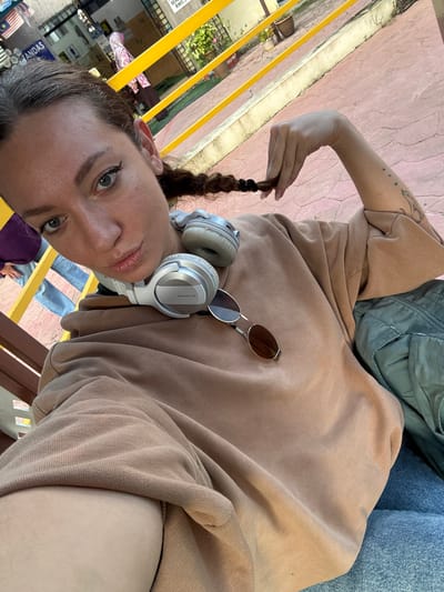 In Wang Kelian, Malaysia, a woman with her hair in a braid is captured in a selfie. She is wearing a brown hoodie and blue jeans, with a pair of white Pioneer DJ headphones around her neck and sunglasses perched on her hoodie. Her left arm is raised, with her hand playfully holding the end of her braid. A subtle tattoo of stars and a cat is visible on her forearm. In the background, a yellow railing divides the scene, with glimpses of a building, some potted plants, and other individuals, one of whom is wearing a pink headscarf and a traditional outfit. The ground appears to be paved with pinkish-red tiles.