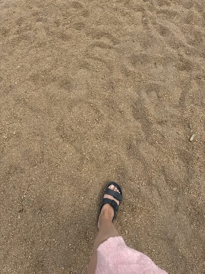 A person's left foot wearing a black sandal and a portion of a pink skirt is visible in the lower left quadrant of the frame, standing on a surface of small, light brown pebbles and sand. The surface appears to be a path or ground in an outdoor setting, possibly within the city of Shalkar, Kazakhstan, characterized by its dry, arid terrain. The lighting suggests it is daytime, with subtle shadows indicating an overcast sky or diffused sunlight.