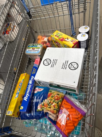 The image captures a shopping cart filled with groceries and household items, likely in a supermarket in Worcester, United States. The cart is viewed from above, looking down into its contents.

The main subjects are various products. Prominently displayed is a white carton of eggs, labeled "CAGE FREE LARGE WHITE EGGS" and "GRADE A EGGS." This carton is placed on its side, despite visible warnings stating "DO NOT PLACE BOX ON ITS SIDE" accompanied by an icon of a box with a diagonal line through it, repeated twice.

Other items include a package of "Organic BABY-CUT CARROTS" and a bag of "BIRDS EYE Mixed Vegetables." Below these, several plastic water bottles are visible. To the left of the egg carton, a yellow box of "MOTTS" applesauce is present, and below it, a clear container of cut fruit or melons. A container of "COUNTRY CROCK" spread, noting "CALCIUM," is also in the cart.

Household goods consist of a blue box of "Reynolds Wrap HEAVY DUTY" aluminum foil and a yellow box of "GLAD Cling 'n Seal" plastic wrap. Two small containers with white caps, possibly salt and pepper shakers, are clipped to the side of the cart. The cart itself has a blue strap with "Walmart" visible on it, indicating the shopping location. The lower shelf of the cart appears to hold white paper plates or similar disposable items.

The scene depicts the aftermath of or an ongoing grocery shopping trip, with various essentials accumulated in the cart. No people are visible. The lighting suggests an indoor setting, typical of a store environment.