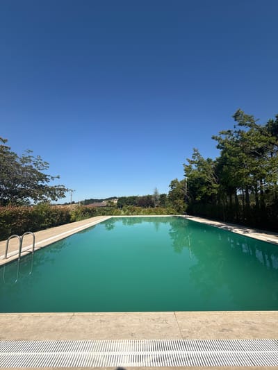 The image shows a swimming pool in Termal, Turkey. The pool is filled with turquoise water and is surrounded by a concrete deck. There are trees and bushes around the pool, and in the background, there are houses and hills. The sky is clear and blue. The image is taken from a low angle, looking across the pool towards the horizon. The pool appears to be empty, and there are no people in the image.