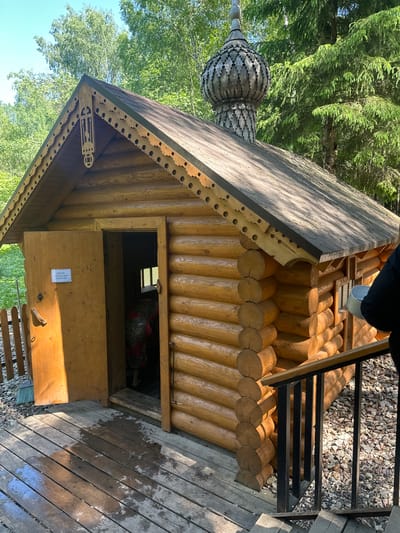 The image depicts a small, wooden, log-built structure, likely a chapel or similar religious building. It features a distinctive, onion-shaped dome characteristic of Russian Orthodox architecture. The structure has a single door that is slightly ajar, revealing a glimpse of the interior. A sign is visible on the door. The surrounding environment suggests a forested area, consistent with a rural location. The presence of a wooden walkway and metal railings indicate that the site is accessible and maintained for visitors. The location is specified as Neglovo, Russia.