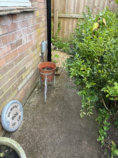 The image depicts an outdoor scene in Longden, United Kingdom. A brick wall runs along the left side of the frame. Adjacent to the wall is a paved area. A brown terracotta pot sits on a metal stand on the pavement. Next to it is a round stone plaque with the inscription "BEWARE OF THE DOG". To the right of the pavement is a green bush. A wooden fence borders the area in the background. A black pipe runs along the top of the brick wall. A cable runs near the wall next to the pipe.