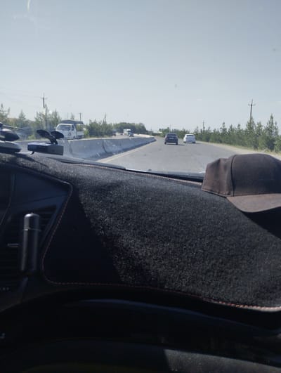 A black baseball cap rests on a textured black dashboard mat in the foreground. Beyond the mat, a windshield reveals a road with a white concrete barrier and several vehicles. A white truck is visible on the road, followed by a black car and a white car. Trees line the right side of the road, and utility poles with wires are scattered along the landscape. The sky is clear and pale blue. The scene appears to be a highway outside of Kattakurgan City, Uzbekistan, possibly during daytime given the bright light.