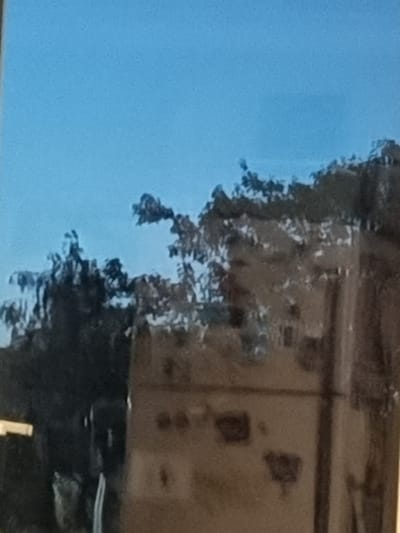 The image depicts a hazy reflection in glass, showing a bright blue sky above a dark, silhouetted treeline. Below the treeline, the reflection of a building with an indistinct facade is visible. The building appears to have windows and other architectural features, but they are obscured by the blurriness of the reflection. The lower part of the image is dominated by the reflection of the building and its surroundings, with some lighter areas that might indicate lights or openings. There are no discernible people or actions taking place. The scene is not clearly identifiable as being in Temernitskiy, Russia, or any other specific location due to the abstract and blurry nature of the reflection.