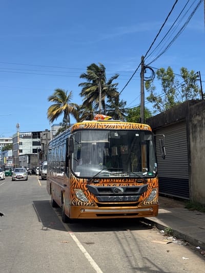An eye-level outdoor shot captures the front and side of a large bus, distinctive for its orange/gold and black tiger-striped livery, parked on the side of a road. The bus's front grille prominently displays the "YUTONG" brand. Its license plate, "SPNU-9248," is visible below the windshield, which also features a small "BSIV" sticker on the upper left. A side mirror extends from the driver's side.
The scene depicts a sunny day in Matara, Sri Lanka, with a clear blue sky overhead. Power lines stretch across the frame, connected to a utility pole. Lush green palm trees and other foliage are scattered throughout, adding to a tropical feel.
To the right of the bus, a concrete sidewalk is visible, showing some debris and litter. In the background, a street extends with other vehicles, including what appears to be a white tuk-tuk and other cars, indicating traffic. Various buildings of different styles and heights line the street, with one showing a corrugated metal facade. Strong sunlight creates shadows on the pavement, highlighting the time of day.
