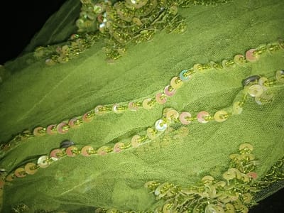 The image displays a close-up view of a vibrant lime-green, sheer, net-like textile, possibly tulle or mesh. The fabric is heavily embellished with numerous small, circular, iridescent sequins, reflecting hues of pale pink, yellow, and green. These sequins are interconnected by strings of tiny, spherical green beads, forming distinct, curvilinear lines and clusters across the surface. At least two small, dark, metallic-appearing fasteners or clips are visible embedded within the beaded sequences, particularly towards the right side of the frame. The lighting highlights the reflective properties of the sequins, creating varying degrees of shimmer. No individuals are present, and no actions or interactions are depicted. The background is indistinct, indicating a very shallow depth of field focused solely on the embellished fabric. The specific location in the city of Na Sang, Laos, cannot be determined from the visual evidence provided in this close-up image of a textile.