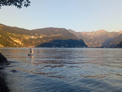 A person is paddleboarding on a calm lake with mountains in the background. The sun is setting, casting a warm glow on the scene. The person is standing on a paddleboard in the middle of the lake, facing away from the camera. They are wearing blue shorts. The lake is calm, with gentle ripples on the surface. The mountains in the background are covered in trees, and there are some buildings visible on the slopes. The sky is clear and blue. The location is Hergiswil (NW), Switzerland.