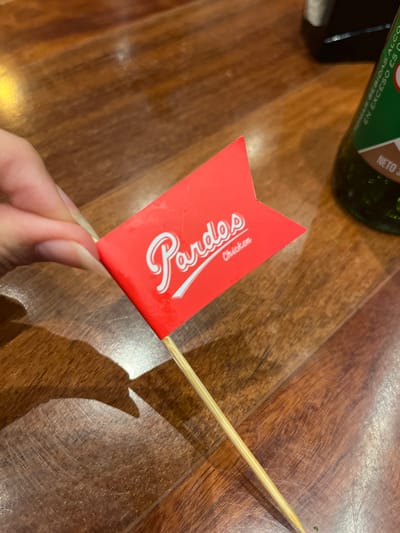 A person's hand is holding a small red flag with a white logo that says "Pardos Chicken" on a wooden table. In the background, a green bottle of Pisco is visible. This scene likely takes place in a restaurant in the city of Santiago de Surco, Peru, as "Pardos Chicken" is a popular Peruvian chain.