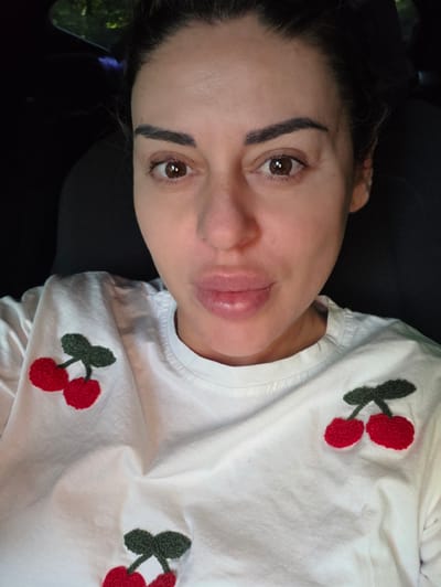 A close-up, frontal, indoor photograph shows a woman with brown eyes and dark hair pulled back. She is wearing a white t-shirt with embroidered red cherries. The woman is looking directly at the camera with her lips slightly parted. The background is dark and out of focus, suggesting an indoor environment. There is no visible indication of the city of Ravnogor, Bulgaria.