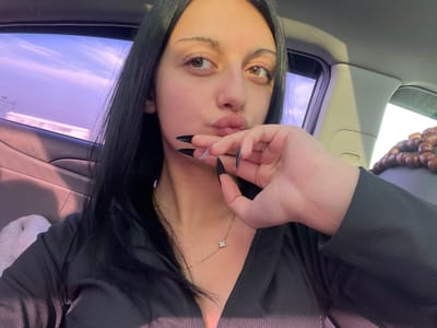 The image portrays a female subject inside a vehicle, presumably located in Akhalsopeli, Georgia. The subject, viewed from the chest up, has long dark hair and brown eyes, and is making a pouting expression with her lips. Her right hand, featuring black, sharply pointed fingernails with clear tips, is positioned near her chin and mouth. She is wearing a black, ribbed, V-neck top and a gold chain necklace with a small, four-petal clover-shaped pendant. The car's interior shows a light-colored fabric surface, likely a seat or headrest, on the left, and a darker door panel. Through the car window on the left, a sky with purple-pink hues and clouds is visible, along with a faint Hyundai logo on the glass. On the upper right side of the image, a string of brown wooden beads is visible, draped over an unseen car fixture.