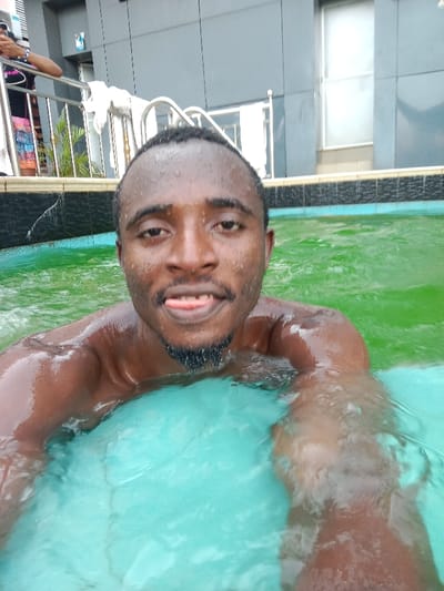 A man is captured in a close-up selfie while swimming in a pool. He appears to be of African descent, with dark skin, short dark hair, and a neatly trimmed goatee. Water droplets are visible on his forehead and hair, indicating he is wet from being in the pool. He is looking directly at the camera with a playful or relaxed expression, his tongue slightly sticking out.

His bare upper body is visible, with his shoulders and arms submerged in the water. The pool water itself is a distinct green-blue hue, with ripples on its surface. Directly in front of him, beneath the water, a lighter blue area, possibly part of the pool's bottom or a floating object, is visible.

In the background, behind the man and to his left, part of another person's arm and torso can be seen behind a silver metal railing. This person appears to be wearing a black top over a colorful patterned garment. A white towel is draped over a section of the railing. Beyond the railing, green foliage is visible. The background also features a modern building with gray, possibly paneled or tiled, walls and what appears to be a large window. A stainless steel pool ladder with white steps is also partially visible in the pool near the railing. The lighting suggests it is daytime.