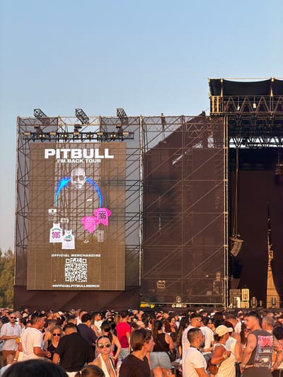 The image captures an outdoor concert scene, likely in Mosta, Malta, during daylight. Dominating the frame is a large stage structure with a massive screen displaying a promotional image for the "PITBULL I'M BACK TOUR." The screen features a portrait of Pitbull, along with graphics of merchandise like jerseys and t-shirts, and a QR code for official merchandise. A large crowd of people is gathered in front of the stage, suggesting they are attending the concert. The environment appears to be an open area, possibly a large field or plaza, with some trees visible in the background. The overall lighting indicates it is daytime, with the sun casting shadows and illuminating the scene.