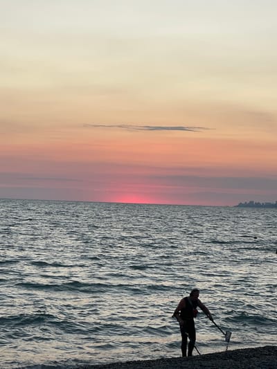 A lone individual wearing dark clothing and a red vest is wading in the shallow water at the edge of a pebble beach, holding a long-handled net. The individual appears to be searching the water or the seabed. The sun is setting over a calm sea, casting warm hues of pink and yellow across the sky. A distant city skyline is visible on the horizon to the right. The water has small waves. The scene is the coast of Krayevsko-Armyanskoye, Russia.
