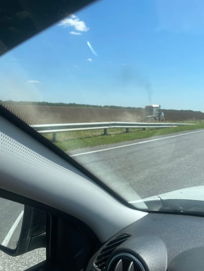 This media file shows a view from inside a vehicle, looking out through the windshield. The scene is a rural landscape with a road and a field. The image is oriented vertically, making it appear as though the viewer is looking sideways at the scene.

The primary subjects are the landscape and the road. A tractor is visible in the field, raising dust as it operates. The sky is blue with a few scattered clouds, suggesting it is daytime and likely a clear day. The road appears to be a highway with guardrails. Inside the vehicle, parts of the dashboard and windshield frame are visible, along with a side mirror.

The activity taking place is agricultural work, with the tractor tilling or spraying the field. The overall impression is one of a bright, sunny day in a rural setting. The presence of the road and vehicle indicates travel through this area.

There is no visible text in the image. The location is identified as Kruchenaya, Russia.