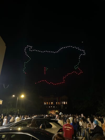 At night in Bratsigovo, Bulgaria, a crowd gathers along a street, some standing beside parked cars, to observe a drone light show in the dark sky. The drones are illuminated in white, green, and red lights, forming a shape reminiscent of the Bulgarian flag. In the background, a building with lit windows can be seen, as well as streetlights illuminating the area.