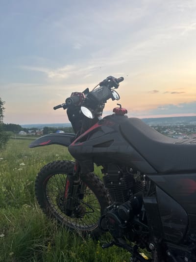 The image shows a dirt bike parked in a grassy field at sunset, with a village and hills visible in the background. The sky is painted with warm hues of orange and yellow, indicative of the setting sun. The dirt bike is black with red accents and features knobby tires, suggesting it's designed for off-road use. The handlebars, mirrors, and a portion of the seat are visible. The rear wheel and part of the engine are also in focus. The grass around the bike appears tall and somewhat overgrown, with some wildflowers interspersed. The scene evokes a sense of peacefulness and adventure, perhaps after a day of riding. The location is identified as Sharan, Russia. The time of day is sunset. There is no visible text in the image.