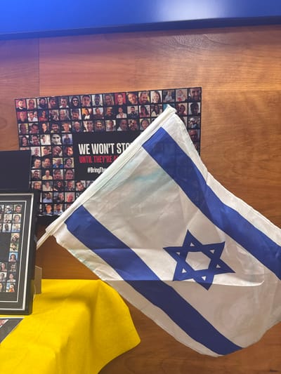 The image displays an indoor scene in the city of Azor, Israel, featuring a patriotic and commemorative display. A white Israeli flag with two horizontal blue stripes and a blue Star of David is prominently positioned in the foreground, draped and slightly unfurled towards the right. A faint light blue discoloration is visible on the flag's upper left white section. In the background, against a light brown wooden paneled wall, a large black poster or board is visible. This poster features a grid of numerous individual portrait photographs of various individuals. Towards the bottom center of this poster, white text on a black background reads "WE WON'T STOP" and below it, "UNTIL THEY'RE A" followed by "#BringThem," strongly suggesting a reference to the "Bring Them Home Now" campaign for Israeli hostages. To the left of this main poster, a smaller black-framed object, also appearing to be a collage of portrait photographs, rests on a surface. A yellow fabric, possibly a tablecloth or drape, is visible in the bottom left foreground, partially covering a surface beneath the framed object and flag. A dark blue strip, possibly a shelf or part of the wall, runs along the top edge of the wooden paneling. No people are actively present in the immediate scene, but their images are central to the posters.