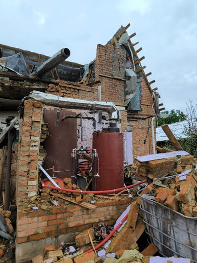 The image displays the extensively damaged remains of a brick building in Pysarivka, Ukraine, under an overcast sky. The structure's roof and upper section have collapsed, revealing the interior. A large, bent metal pipe, possibly a chimney or vent, protrudes from the wreckage of the upper left wall. Within the exposed building section, two large, reddish-brown cylindrical tanks, connected by various black and red pipes, valves, and gauges, are visible. The ground surrounding the building and extending into the foreground is covered with a substantial accumulation of debris, consisting primarily of broken red bricks, splintered wood, fragments of white and light purple insulation material, and other building components. A large metal cage, partially filled with rubble, is situated in the lower right foreground. Green foliage is visible beyond the damaged structure on the right, with another building faintly discernible in the distant background. No individuals are present in the scene.