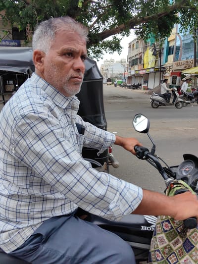 The image depicts a man with grey hair and a beard, wearing a blue and white checkered shirt and dark pants, sitting on a black motorcycle. He is holding the handlebars, with his right hand on the throttle. His gaze is directed to the right, and his expression is serious. The motorcycle is parked on a paved road, with other motorcycles and an auto-rickshaw visible in the background. Buildings line the street, some with shops on the ground floor. A large tree with green leaves provides shade over the scene. The environment suggests a street in a city, possibly Datala, India, given the context. The lighting indicates it is daytime, and the overall atmosphere is casual, likely a typical street scene in India.