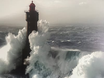 A tall, dark lighthouse with a red cap stands in the foreground, battered by colossal waves. White foam erupts from the impact of the water against the structure, obscuring much of its lower half. The sea is rough, with dark, choppy water stretching to the horizon under a pale, hazy sky. The scene evokes a sense of powerful natural forces at play. There is no indication of Sherbakul, Russia, in this image; it is a coastal scene.