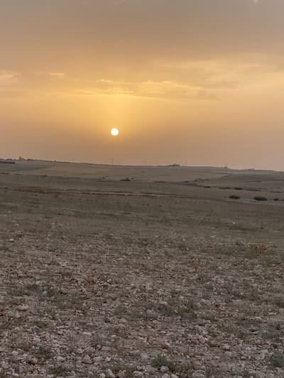 The image captures a landscape scene in Agafay, Morocco, during what appears to be sunset or sunrise. The sky is a soft, peachy-orange hue, with diffused sunlight visible as a bright, white orb near the center. Wispy clouds are scattered across the upper portion of the sky.

The terrain below is characterized by rolling hills covered in dry, sparse vegetation and scattered rocks. The foreground is dominated by a textured ground surface composed of numerous light-colored stones and pebbles, interspersed with dry grass and small plants. In the mid-ground, the landscape transitions to fields that appear more cultivated or naturally sparse, with patches of darker green vegetation visible. Further in the distance, along the horizon, a series of structures and utility poles are discernible, suggesting the presence of a settlement or infrastructure. The overall atmosphere is serene and evokes a sense of open, arid land.