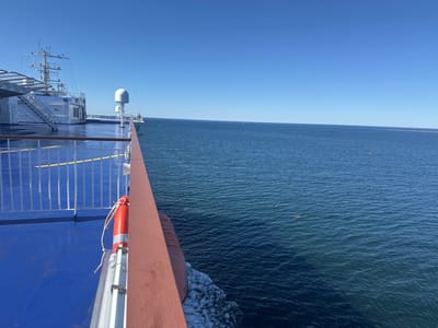 An outdoor, eye-level, wide shot depicts the deck of a ferry on a clear, sunny day. The ferry is moving, as indicated by the white wake trailing from its side. The ferry's blue deck occupies the left side of the frame, with a white railing and a wooden railing framing the view. A bright orange life preserver is attached to the railing. In the distance, the calm, deep blue sea stretches to the horizon, meeting a clear blue sky. The mast and superstructure of the ferry are visible on the left side, with various antennas and equipment. The distant horizon shows faint outlines of land or structures. A small, dark object, possibly a buoy, is visible in the water to the right of the frame. The scene suggests a calm sea voyage.
