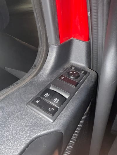 The image shows a close-up of a car's interior door panel. Dominating the frame are a set of black plastic buttons and controls. These include a directional pad with four arrows, likely for adjusting mirrors, and several other buttons with icons. To the left, there are two rectangular buttons, one with a lock icon and another with an unlock icon. Above the directional pad, there are two more rectangular buttons, one with a camera-like icon and another with a fold-out mirror icon. The car's interior appears to be black and red. The image is taken from a slightly elevated angle, looking down at the controls. There is no indication of people, actions, or interactions. The environment is limited to the car's interior. The location is stated to be the city of Wildwood, United States.