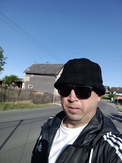 The image is a selfie taken by a man wearing a black bucket hat and sunglasses. He is also wearing a black and white Adidas jacket over a white t-shirt. The man is standing on a paved road in what appears to be a rural or semi-rural area. In the background, there is a fence, a house with a grey facade and a dark roof, and trees. The sky is clear and blue. The location is specified as Čistá, Czechia.