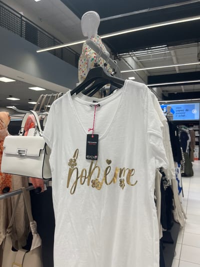 The image shows a white t-shirt with a gold "bohème" inscription on a hanger inside a clothing store. The store is located in Saint-Brice-Courcelles, France, at coordinates 49.283 latitude and 4 longitude. There is a white purse hanging next to the t-shirt. In the background, a mannequin is wearing a multi-colored shirt. Several other garments hang on hangers in the background. The clothing store appears to be well-lit and well-organized. The image does not include any people. A sign in French is visible in the background advertising a potential prize for a purchase. The t-shirt has a small pink tag hanging from it.