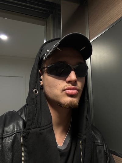 The scene depicts a close-up portrait of an individual inside a building located in Asnières-sur-Seine, France. The subject, appearing to be a young male, is seen from the chest up, facing slightly right with a neutral expression and lips slightly parted. He wears a black baseball cap adorned with four silver-colored studs or spikes on the brim, dark sunglasses (Bolle brand visible on the left temple), a black hooded sweatshirt with frayed edges and a silver grommet on the left side of the hood, and a black leather jacket with a partially visible zipper. A gray undershirt is visible beneath the hoodie. A light brown goatee and mustache are present. A silver-colored hoop earring is visible in the subject's left ear. The background on the left consists of a white wall with a recessed light fixture and a metallic vertical beam. On the right, dark gray or black paneling is visible, partially obscured by a diagonally oriented wooden-textured panel above. The reflections in the sunglasses suggest an indoor, possibly well-lit, environment.