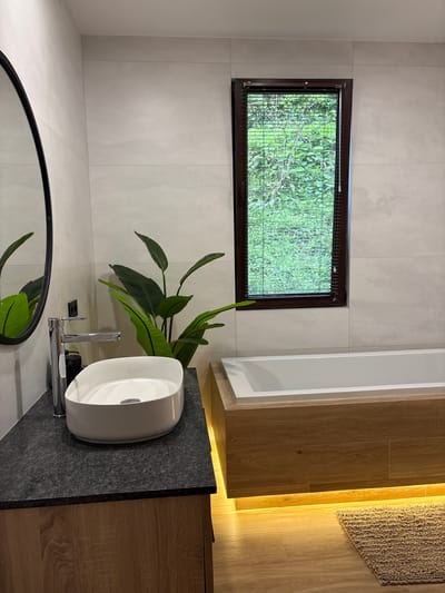 The image depicts a bathroom in what is likely a private residence in Khala, Georgia. The walls are covered in large, light gray tiles. A rectangular window with a dark brown frame sits high on the wall, offering a view of lush greenery outside, obscured by a screen or blinds. Below the window, a white vessel sink is mounted on a dark gray countertop. A chrome faucet is installed on the countertop next to the sink. A tall, leafy plant stands behind the sink, partially obscuring the window. A round mirror with a black frame is visible at the bottom of the image, reflecting the plant. To the right of the sink, wooden cabinets with a light brown wood grain pattern are visible. A light-colored bathmat is on the floor, partially visible in the upper right corner of the image. There are no people visible in the scene.