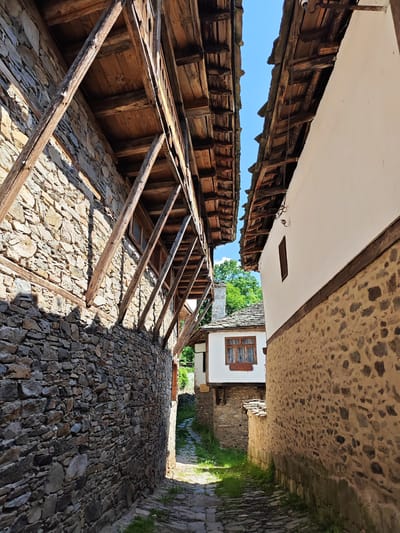 A narrow, stone-paved alleyway winds between traditional houses in the village of Kovachevitsa, Bulgaria, under a clear blue sky. On the left, a multi-story structure features a lower wall of rough-hewn, irregular grey and brown stones. Above this, a prominent wooden overhang is supported by multiple diagonal dark brown wooden beams, creating a patterned shadow on the stone wall. The underside of the overhang reveals horizontal wooden planks. A series of small, dark rectangular windows are visible beneath the wooden soffit.
On the right, a house exhibits a lower wall of lighter, more uniformly textured stone, topped by a smooth, white plastered upper wall. A dark horizontal wooden beam separates the stone from the plaster. A single, small, closed, dark wooden window is set into the white wall. The roofline features traditional dark stone slabs.
The cobblestone path is uneven, with patches of green grass growing between the stones. Further down the alley, a distant white house with a prominent multi-pane wooden window is visible, with a sign below it reading "КЪЩА-МУЗЕЙ / КОВАЧЕВИЦА" (HOUSE-MUSEUM / KOVACHEVITSA). A stone chimney rises from this distant house. Lush green trees are visible beyond the structures. The scene is illuminated by bright sunlight, casting strong shadows and highlighting textures. No individuals or animals are present.