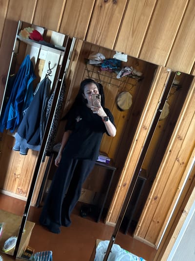 A woman is taking a full-body mirror selfie in a room with wooden paneled walls. She is wearing a black t-shirt and black pants. She is holding a phone in her right hand, taking the picture. There are several coats hanging on hooks on the wall to her left. There are shelves above the hooks holding various items, including a red and yellow hat. To her right, the mirror reflects more of the room, including a shelf with items and two circular decorative items. The floor is reddish-brown. The overall impression is a casual, lived-in space.