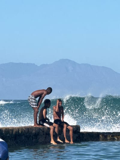 The image shows three young men standing on a weathered stone ledge by the ocean in Muizenberg, South Africa. The man closest to the foreground is Black and wears checkered swim trunks. The other two men wear dark swim attire. A wave is crashing against the ledge, creating white spray. The sky is clear and blue, and a mountain is visible in the background.