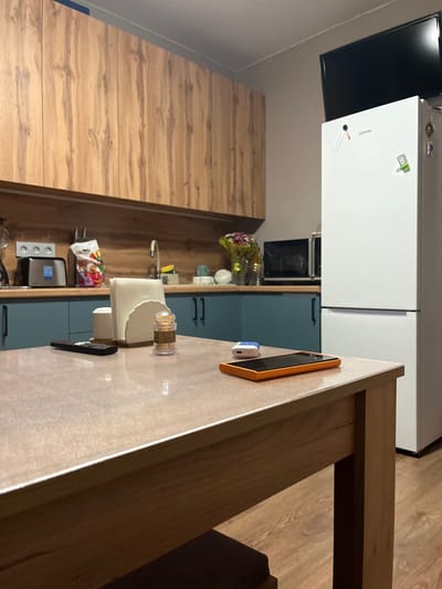This is a medium shot of a kitchen scene, taken from a low angle, with the camera rotated. The foreground features a wooden dining table with a light-colored countertop. On the countertop, there's an orange smartphone and a white wireless earbud case. To the left of these objects, a napkin holder with white napkins and a dispenser of toothpicks are visible. Further left, a bag of colorful snacks is placed on the counter.
In the mid-ground, a white refrigerator stands prominently. On its door, there are several magnets, including what appears to be a calculator and a pen. Below the refrigerator, a built-in oven is visible within blue kitchen cabinets. The cabinets are equipped with black handles. To the far left, wooden kitchen cabinets with a natural wood grain are present.
The background consists of a light-colored wall and what appears to be a wooden floor with a visible grain pattern. The overall setting is a modern kitchen, likely in a residential space. The lighting suggests it is daytime, possibly indoor lighting. There are no people visible in the image. The visible text is limited to the branding on the refrigerator, which appears to be "Gorenje."