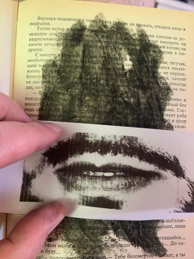 The image is a close-up of an open book with text in Russian. A portion of a black and white drawing or printed image of lips is placed over the text. The book pages are yellowed with age, and a human finger, with a slightly dirty nail, is visible in the lower left corner, holding down the drawing. The text discusses a character named Varvara, who gets up and falls on the bed, and Ustein, who is also mentioned. The drawing of lips is abstract and obscures much of the text, with the lips appearing somewhat smudged. The background appears to be a dark, textured area, possibly part of the drawing or an overlay. There is no visible information in the image that would allow for determining a location in the city of Ganino, Russia.