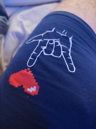 A close-up, high-angle shot shows a navy blue fabric, likely clothing, with two graphic elements printed on it. In the upper portion of the image, a white outline drawing depicts a hand in a "rock on" or "devil horns" gesture, with the index and pinky fingers extended upwards and the thumb and middle fingers curled downwards. Below and to the left of the hand graphic, a red and white pixelated heart shape is visible. The heart is rendered in a retro, video-game aesthetic, with predominantly red pixels and a few white pixels forming details. The fabric has a slight texture, suggesting it is cotton or a similar material. The background is softly blurred, showing a light-colored surface and hints of another fabric or object in a darker shade, creating a shallow depth of field. The image does not contain any elements that would allow for location identification in Grohotno, Bulgaria.