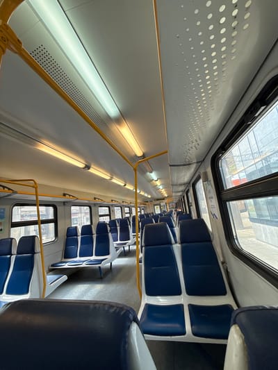 The image shows the interior of a passenger train car in Aprelevka, Russia. The car is empty, with rows of blue and white seats facing forward. The seats are arranged in pairs on either side of a central aisle. Yellow handrails run along the ceiling of the car, and fluorescent lights provide illumination. The windows on the right side of the car offer a view of a train station platform and buildings. The floor of the car appears to be made of a gray, textured material. The overall impression is one of a modern, clean, and functional public transportation vehicle.