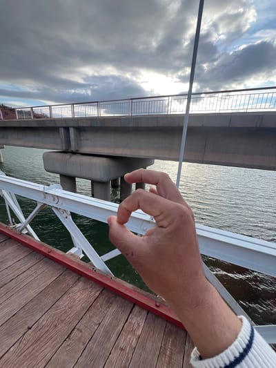 A person's hand is positioned in the foreground, making an "okay" gesture. The background reveals a bridge structure with two levels; the lower level has concrete pillars, while the upper level is steel and concrete. Water is visible beneath the bridge. The sky is overcast with dark clouds. The location is likely a bridge in Tabaroucht, Morocco, suggested by the subject.