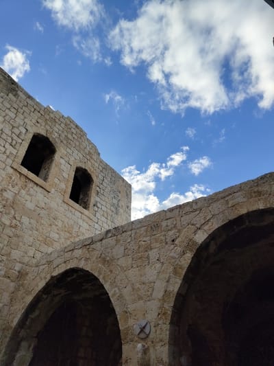 The image captures a low-angle perspective of ancient stone architecture under a partly cloudy blue sky, located in Rosh HaAyin, Israel. The foreground features two large, light-colored stone arches, constructed from irregular blocks. A circular, light-toned object with a debossed cross-like pattern is affixed to the stone wall between the arches. To the left, a taller section of a stone building rises, also made of rough-hewn, light-colored stone blocks. This structure displays two distinct, dark, empty rectangular window openings on its upper facade. The top edge of the building exhibits an uneven, partially ruined appearance. No individuals or active subjects are present in the scene.
