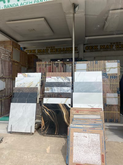 This media file shows a retail display of ceramic tiles in what appears to be a shop. The tiles are stacked in boxes and also displayed as individual samples. Some of the boxes have text indicating brands like "JUPITER," "MARBELLA WHITE," and "GARUDA TILE." Other text on the boxes includes "Keram Kualitas Super," which translates to "Super Quality Ceramics." There are also samples of large format tiles with marble and abstract patterns. Some tiles have text like "CERAMIKA ITALIANA PREMIER" and "OLIVERA." A prominent sign in the background reads "TOKO CEK MAT KERAMIK" with the address "JLN. MEDAN - BANDA ACEH, PANTON LABU - AGEN UTARA," indicating the location is likely in Panton Labu, North Aceh, Indonesia. The lighting suggests it is daytime. There are no people clearly visible in the image, but the setting is clearly a store or warehouse specializing in tiles.