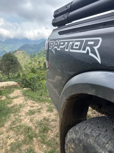 The image shows the rear of a black Ford Raptor truck parked on an unpaved surface overlooking a valley in Suối Giàng, Vietnam. The "RAPTOR" emblem is visible on the rear quarter panel. The truck's tire is visible on the right side. The landscape is characterized by lush greenery and mountain ranges in the distance under an overcast sky. The ground immediately around the truck consists of dirt, grass, and small rocks.