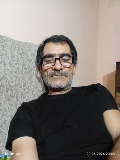 A medium shot shows a middle-aged man with dark hair, glasses, and a mustache, sitting in a chair. He is wearing a black t-shirt. The background is a pale pink wall with a patterned couch on the left. The timestamp "29.06.2026 23:03" and "REDMI A3" are visible in the lower right and lower left corners, respectively. The setting is indoors, possibly a home in Pshap, Abkhazia.
