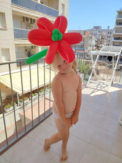 A young girl with blonde hair is standing on a balcony, wearing a red and green balloon flower hat. She is wearing white underwear and her hands are clasped in front of her. She is smiling and looking at the camera. The background shows buildings, a clothes drying rack, and a clear blue sky, indicating it is daytime and likely sunny. The balcony has a tiled floor and a metal railing with a yellow and white striped awning visible below. The presence of air conditioning units on the building in the background and the architecture suggest a warm climate, consistent with the location of Calafell, Spain. The overall mood is playful and cheerful, with the child's pose and expression contributing to this feeling.