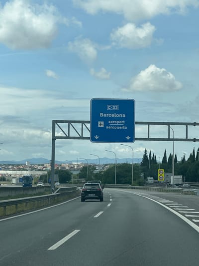 The image captures a daytime scene on a multi-lane highway, viewed from a moving vehicle. A large, blue overhead highway sign is prominently featured in the midground, indicating directions. The sign displays "C-33" at the top, followed by "Barcelona" and "aeroport" (Catalan for airport) with an airplane icon, and below that "aeropuerto" (Spanish for airport). White arrows on the sign point towards the right, indicating the direction for these destinations. The sign is supported by a gray metal gantry structure spanning across the road.
To the right of the highway, further in the distance, a yellow rectangular sign with a red border displays the number "100" and a truck pictogram, likely indicating a speed limit for trucks or specific lane usage. The highway itself is asphalt with clear white lane markings and a white solid line on the shoulder. It curves gently to the right. A black car is visible in the rightmost lane.
Along the right side of the highway, there's a metal guardrail, followed by a strip of grass and a line of tall conifer trees. Beyond these trees, a landscape of low mountains or hills is visible under a partly cloudy sky. More vehicles, including trucks and a blue bus, can be seen on the highway and what appears to be a lower road structure in the background. The sky above is bright blue with scattered white clouds. The setting is a clear day, indicating good visibility. Based on the visible text and context, the location is a highway in Spain, likely near Montmeló, leading towards Barcelona and its airport.